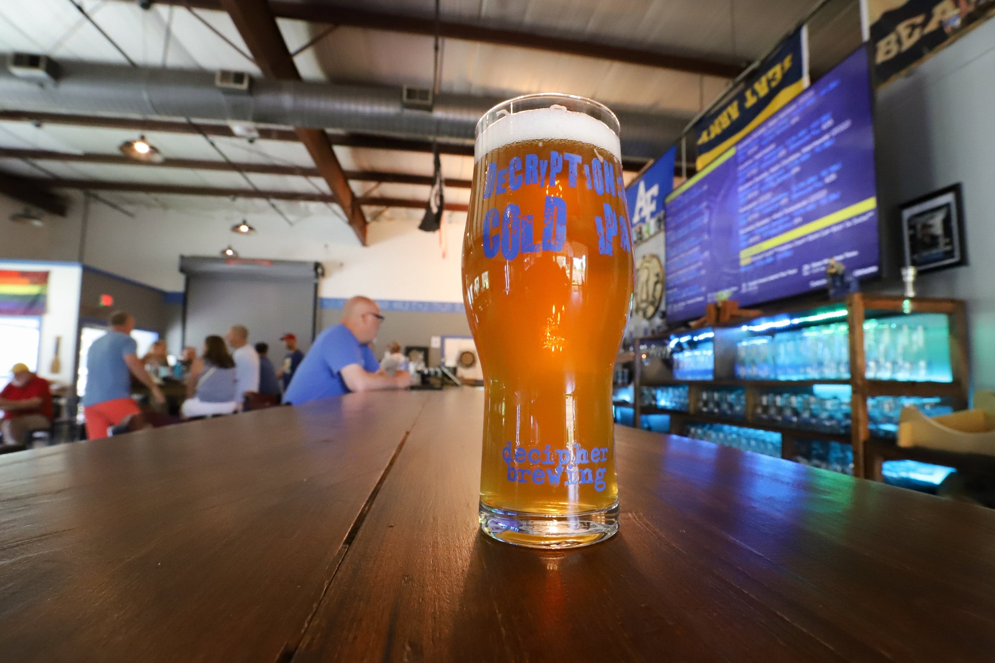 Glass of beer with 'Decipher' branding on a bar counter with people in the background
