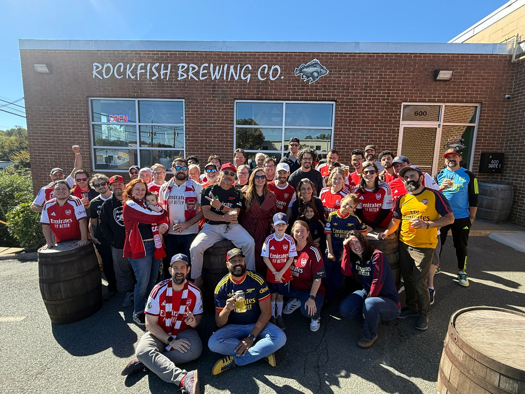 A large group of people posing together outdoors in front of a brick building labeled 'ROCKFISH BREWING CO.' Several people are wearing red and white patterned shirts and holding beer glasses, with barrels and the brewery entrance visible in the background.