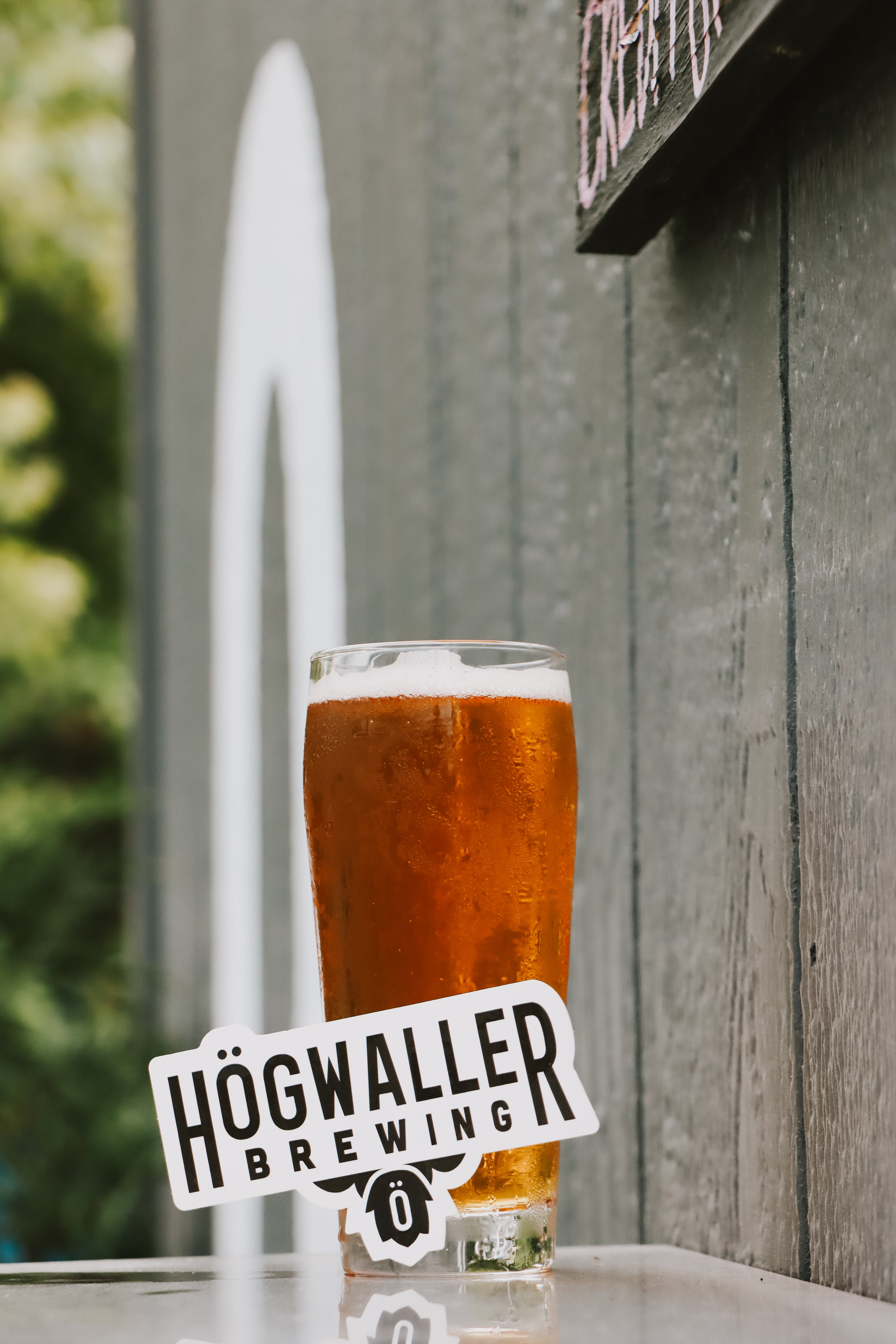 A clear pint glass filled with amber beer and topped with white foam, set on a table in front of a gray wooden wall. A white Högwaller Brewing logo sign is placed in front of the glass.