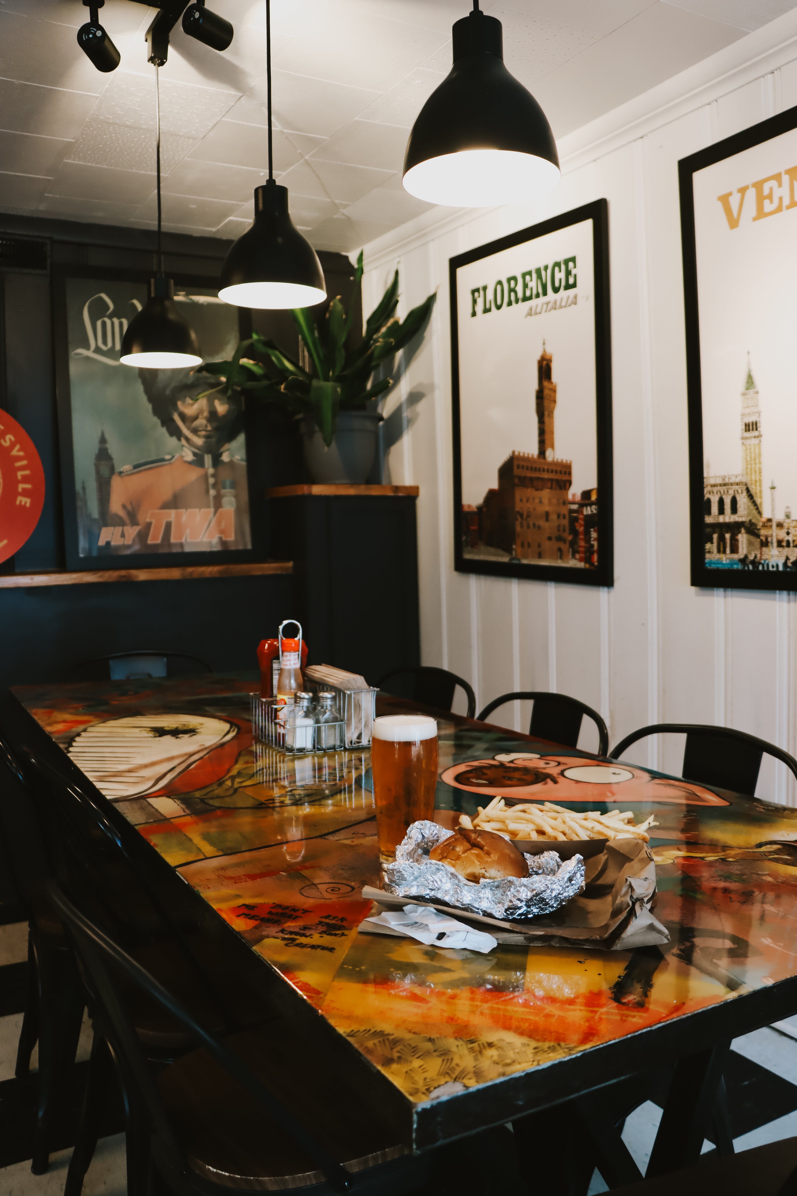 A rustic dining table with a glossy, colorful distressed print featuring orange, yellow, and red tones, set in a restaurant or cafe with black pendant lights, chairs, and wall art.