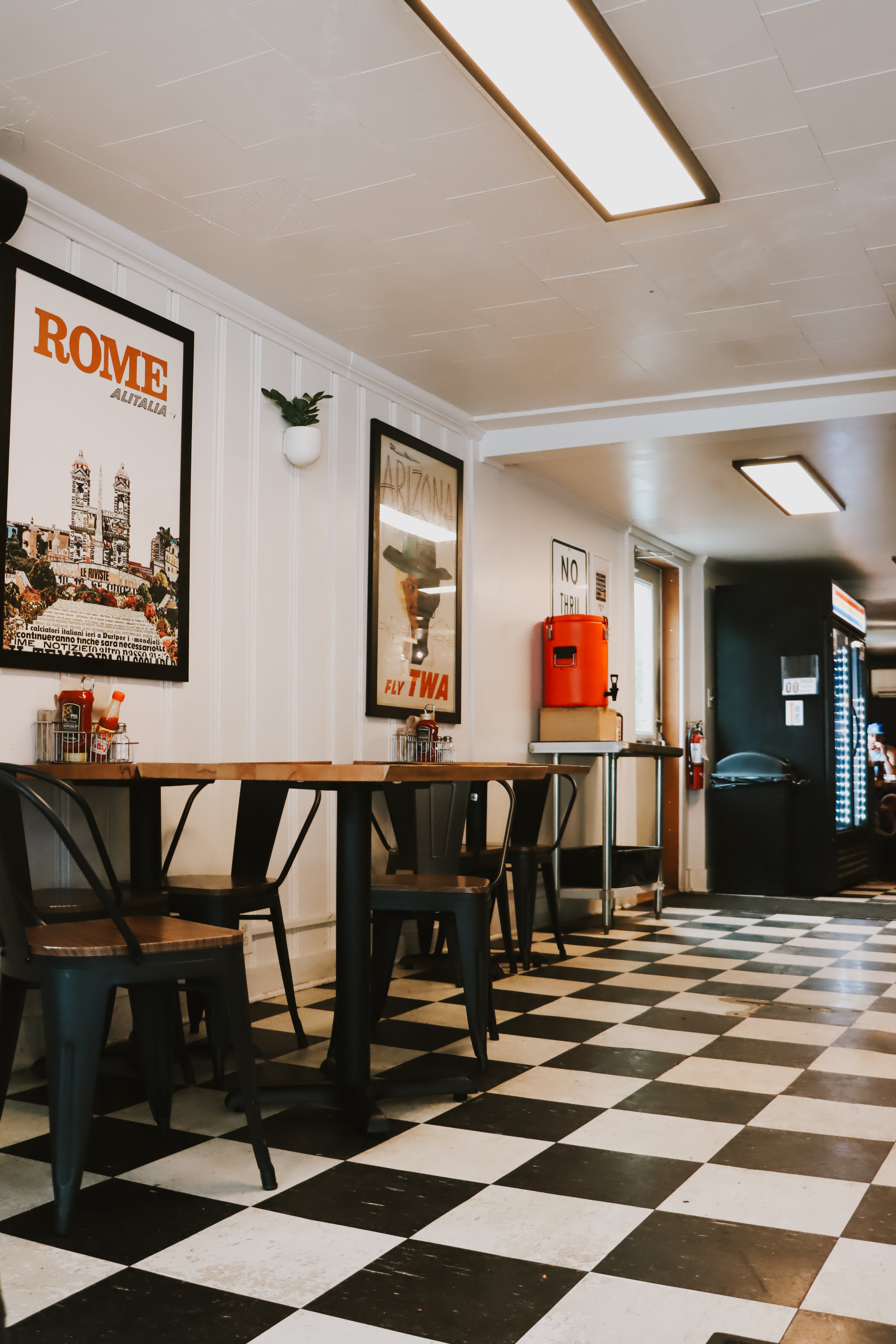 A retro-style restaurant or diner interior with a black and white checkered floor, wooden tables, black metal chairs, framed Rome and New York posters on the white walls, and a bright fluorescent light overhead.