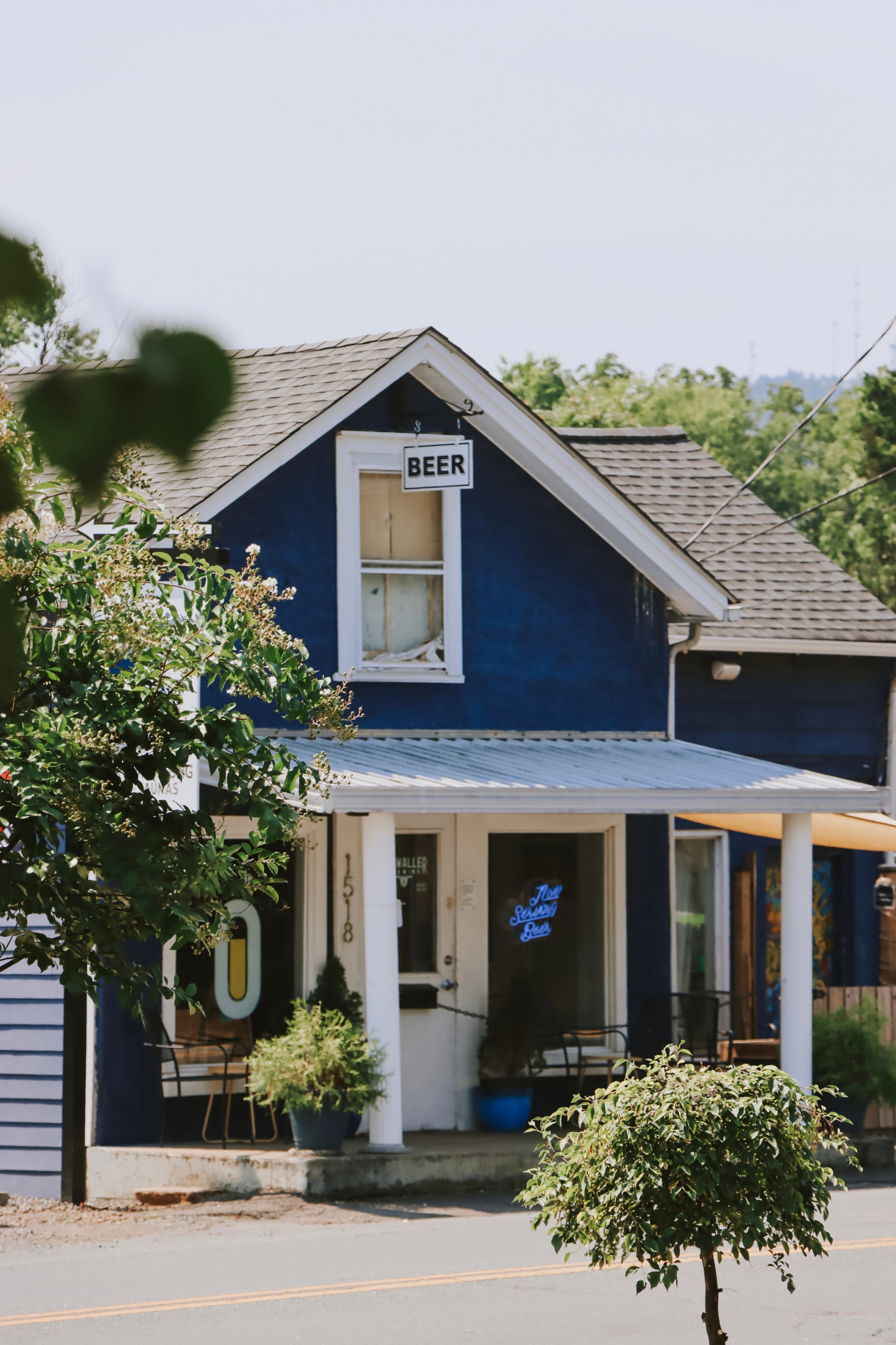 A blue house with a front porch and a hanging 'BEER' sign, photographed outdoors with trees, street, and a clear sky in the background.