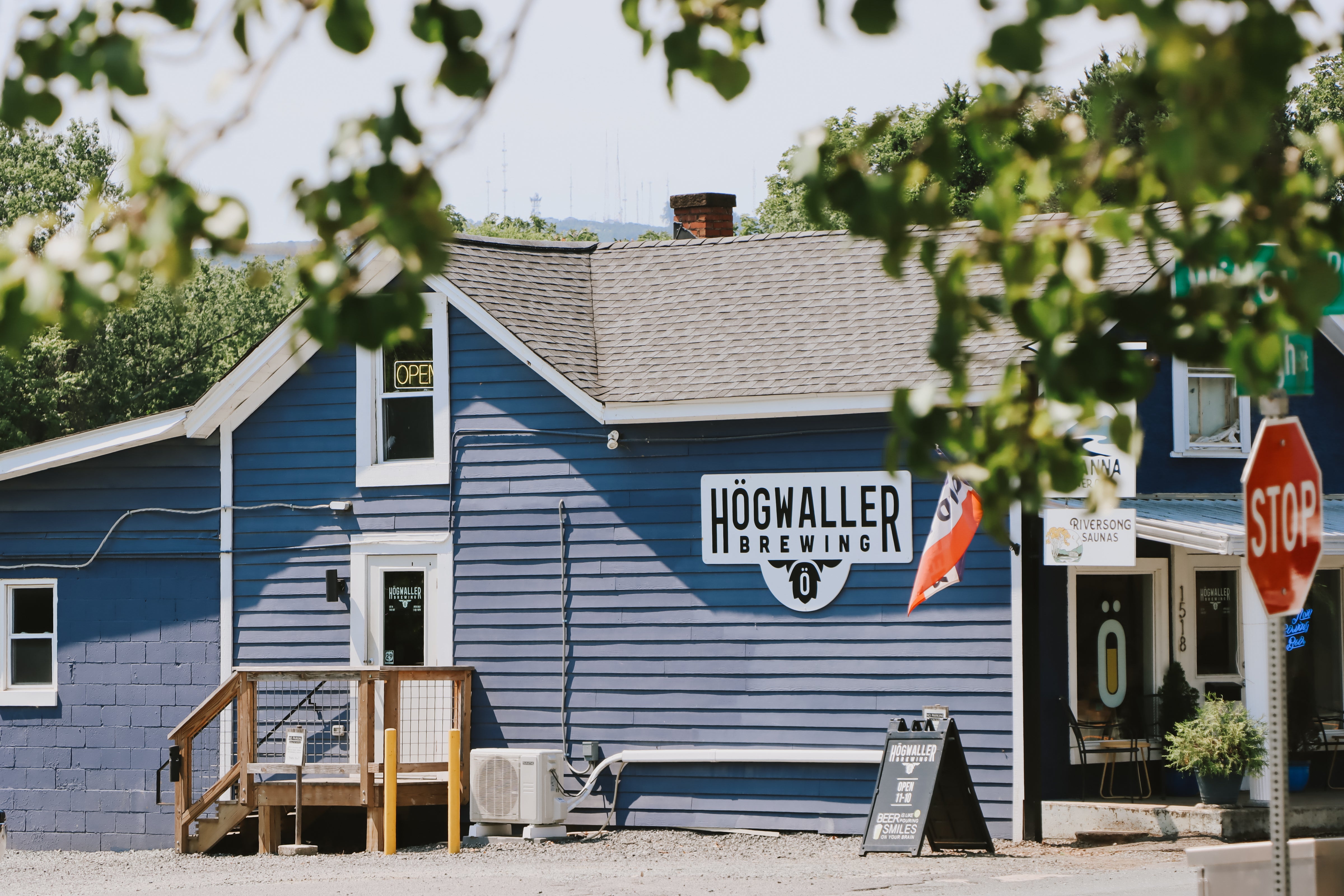 A blue wooden building with a large white sign reading HöGWALLER BREWING, shown outdoors with trees in the foreground and a stop sign on the right.