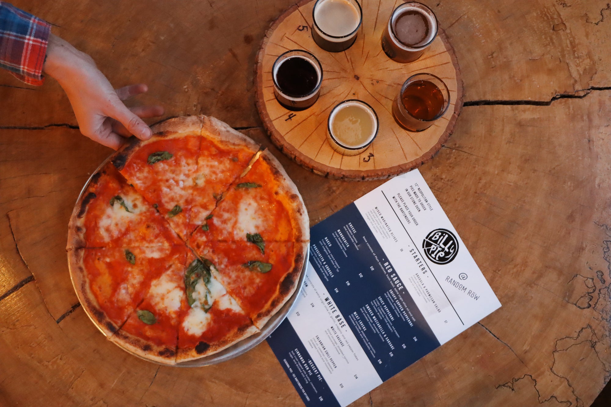 Hand holding a pizza on a wooden table with drinks and a menu.