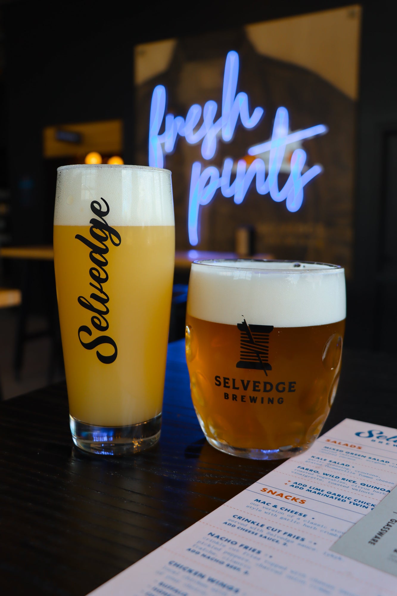 Two glasses of beer from Selvedge Brewing with a neon sign in the background.