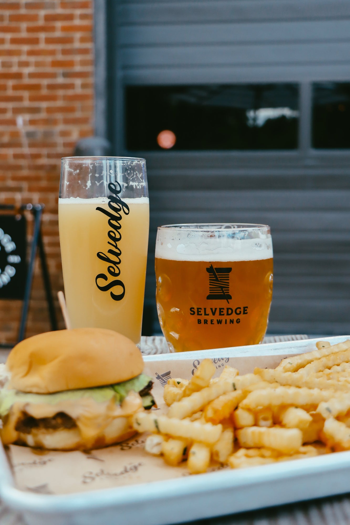 Tray with a burger, fries, and two glasses of beer from Selvedge Brewing on a blurred outdoor background.