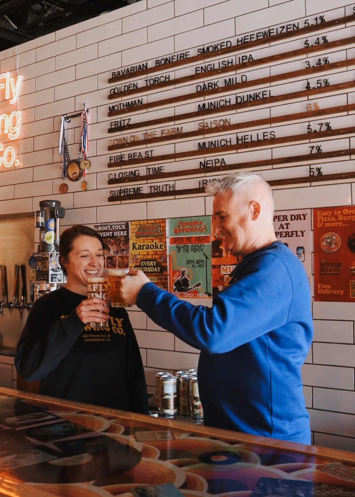 Two people at a bar, one holding a beer, with a beer menu on the wall.