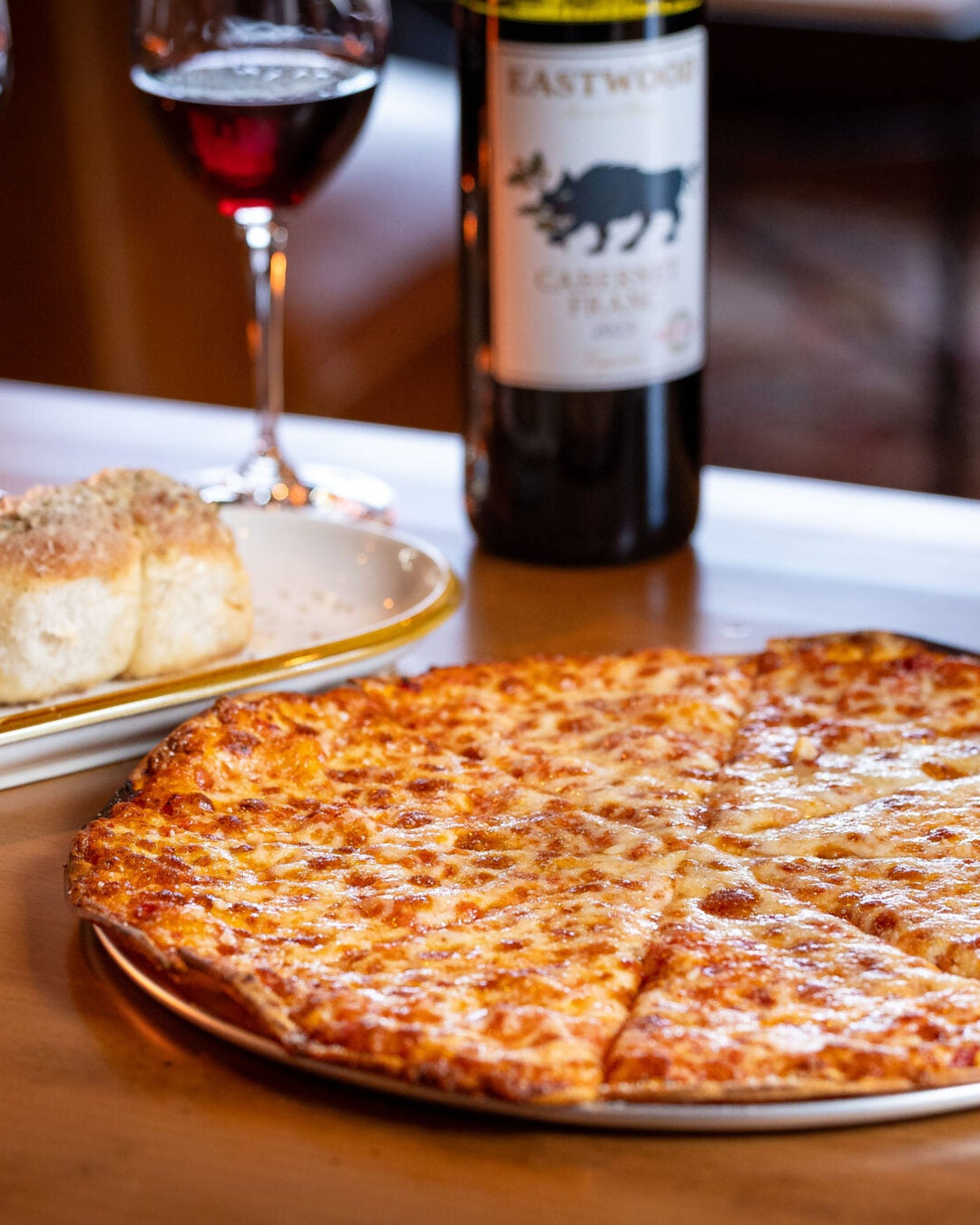Cheese pizza on a table with wine and bread rolls in the background