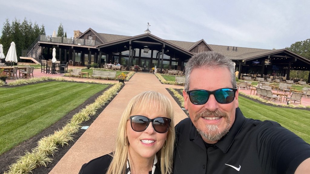 A man and a woman taking a selfie in front of a large lodge-style building with manicured green lawns and a paved walkway. The man is wearing a black Nike polo shirt and sunglasses, and the woman is wearing a black top and sunglasses.
