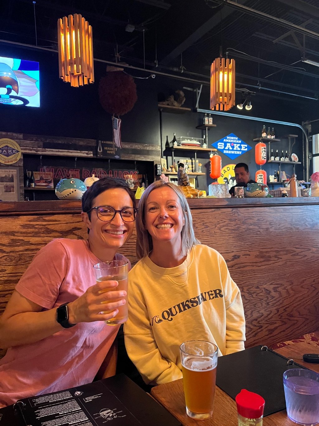Two women smiling at a restaurant booth, with one wearing a pink t-shirt and the other wearing a yellow Quiksilver sweatshirt. A pint of beer and a glass are on the table in front of them, with a bar visible in the background.